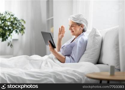 technology, old age and people concept - senior woman with tablet pc computer and wireless earphones having video call in bed at home bedroom. old woman with tablet pc having video call in bed