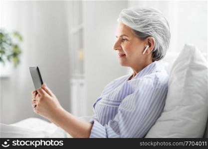 technology, old age and people concept - senior woman with tablet pc computer and wireless earphones in bed at home bedroom. senior woman with tablet pc and earphones in bed