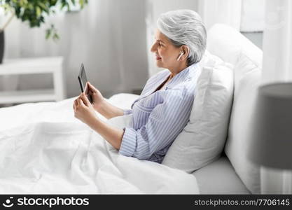 technology, old age and people concept - senior woman with tablet pc computer and wireless earphones in bed at home bedroom. senior woman with tablet pc and earphones in bed