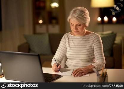 technology, old age and people concept - senior woman writing to notebook with laptop at home in evening. senior woman writing to notebook at home at night