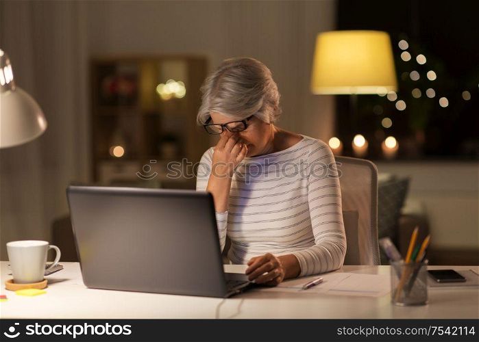 technology, old age and people concept - tired senior woman in glasses with laptop working at home in evening. tired senior woman with laptop at home at night