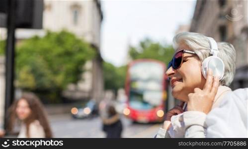 technology, old people and travel concept - senior woman in headphones and sunglasses listening to music over london city background. old woman in headphones listens to music in london