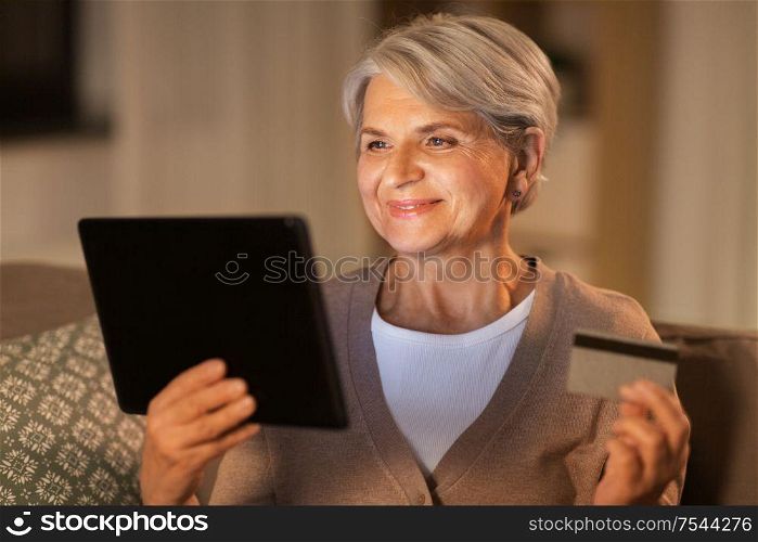 technology, online shopping, age and people concept - happy senior woman with tablet pc computer and credit or bank card at home in evening. old woman with tablet pc and credit card at home
