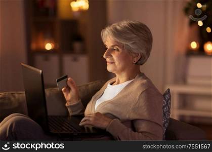 technology, online shopping, age and people concept - happy senior woman with laptop computer and credit or bank card at home in evening. senior woman with laptop and credit card at night