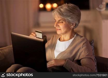 technology, online shopping, age and people concept - happy senior woman with laptop computer and credit or bank card at home in evening. senior woman with laptop and credit card at night
