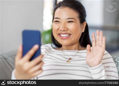 technology, people and communication concept - happy smiling asian woman with smartphone having video call at home and waving hand. asian woman with smartphone having video call