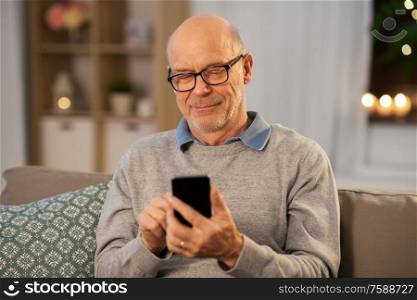 technology, people and communication concept - happy smiling bald senior man texting on smartphone at home in evening. happy senior man texting on smartphone at home
