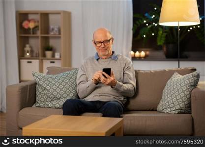 technology, people and communication concept - happy smiling bald senior man texting on smartphone at home in evening. happy senior man texting on smartphone at home