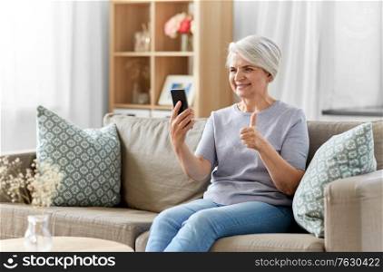 technology, people and communication concept - happy smiling senior woman with smartphone having video call and showing thumbs up at home. senior woman having video call on smartphone