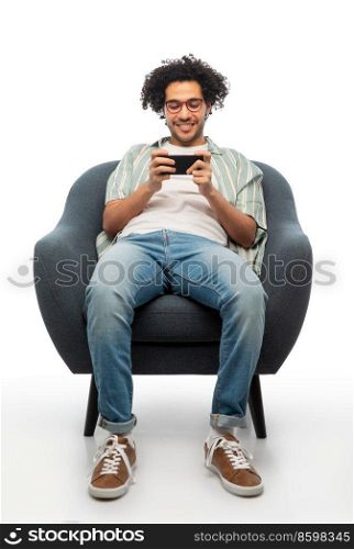 technology, people and furniture concept - happy smiling young man in glasses with smartphone sitting in chair over white background. smiling young man with smartphone sitting in chair