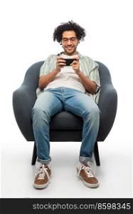 technology, people and furniture concept - happy smiling young man in glasses with smartphone sitting in chair over white background. smiling young man with smartphone sitting in chair