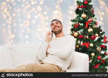 technology, people and holidays concept - smiling man calling on smartphone over christmas tree and lights background