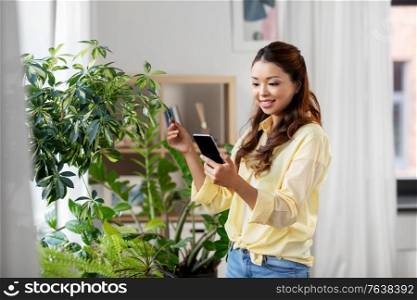 technology, people and internet concept - happy smiling asian woman with smartphone and flowers at home. asian woman with smartphone and flowers at home