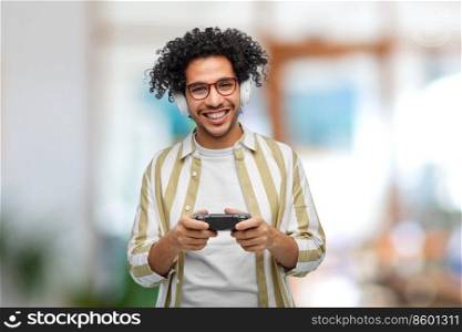 technology, people and leisure concept - happy smiling young man in headphones with gamepad playing video game over office background. man with gamepad playing video game at office