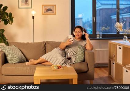 technology, people and leisure concept - happy young woman in headphones listening to music on smartphone at home in evening. woman in headphones listens to music on smartphone