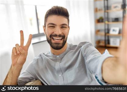 technology, people and lifestyle concept - happy man taking selfie or having video call and showing peace gesture at home. man taking selfie or having video call at home