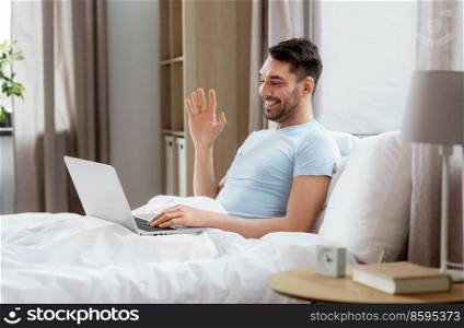 technology, people and lifestyle concept - happy man with laptop computer having video call in bed at home bedroom. man with laptop computer having video call in bed