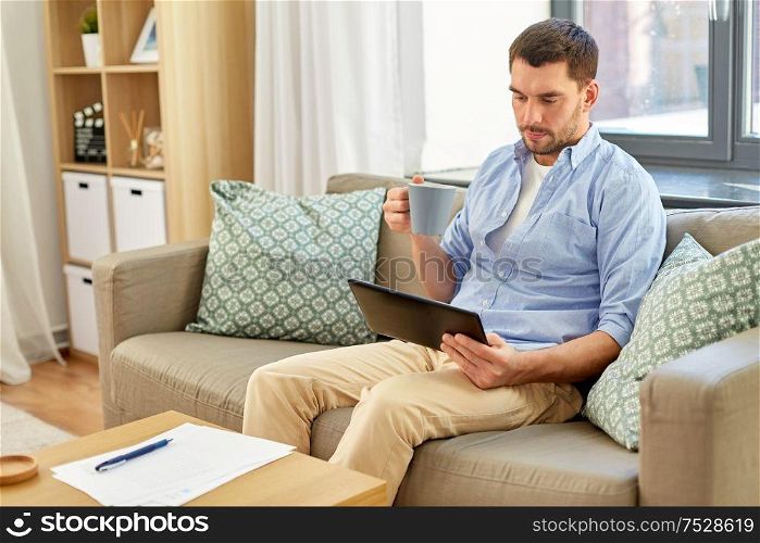 technology, people and lifestyle concept - happy man with tablet pc computer and papers drinking coffee or tea at home. smiling man with tablet pc drinking coffee at home
