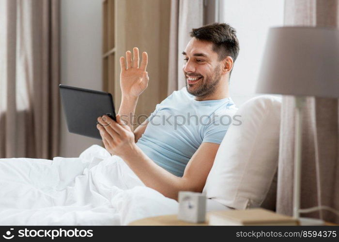 technology, people and lifestyle concept - happy man with tablet pc computer having video call in bed at home bedroom. man with tablet computer having video call in bed