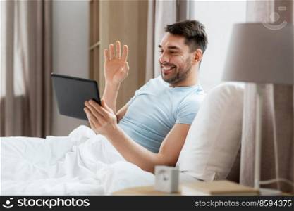 technology, people and lifestyle concept - happy man with tablet pc computer having video call in bed at home bedroom. man with tablet computer having video call in bed