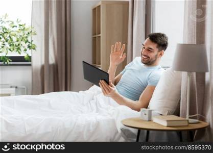 technology, people and lifestyle concept - happy man with tablet pc computer having video call in bed at home bedroom. man with tablet computer having video call in bed