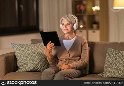 technology, people and lifestyle concept - happy senior woman in headphones and tablet pc computer listening to music at home in evening. senior woman in headphones listening to music
