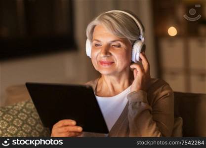 technology, people and lifestyle concept - happy senior woman in headphones and tablet pc computer listening to music at home in evening. senior woman in headphones listening to music