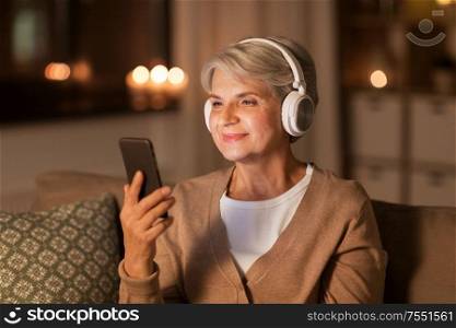 technology, people and lifestyle concept - happy senior woman in headphones listening to music on smartphone at home in evening. senior woman in headphones listening to music
