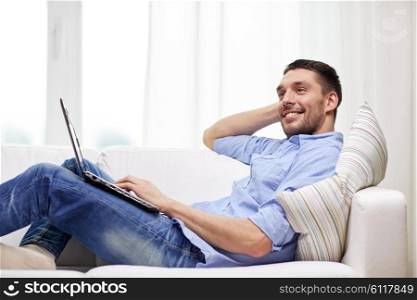 technology, people and lifestyle concept - smiling man lying on couch with laptop computer at home