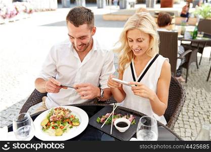 technology, people, eating and leisure concept - happy couple with smatphone taking picture of food at restaurant terrace