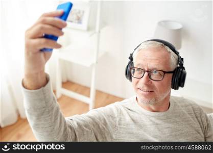 technology, people, lifestyle and leisure concept - senior man in headphones listening to music and taking selfie with smartphone at home