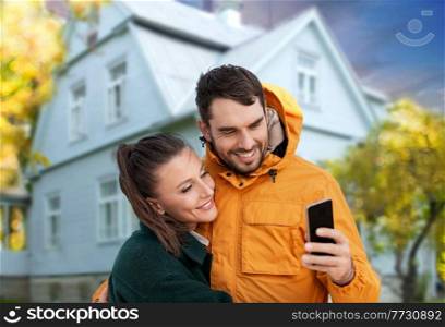 technology, real estate and people concept - happy couple with smartphone taking selfie over house in autumn on background. couple taking selfie over house in autumn