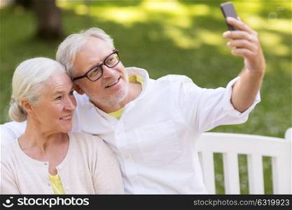 technology, relationship and old people concept - happy senior couple with smartphone taking selfie and hugging at summer park. senior couple taking selfie by smartphone at park
