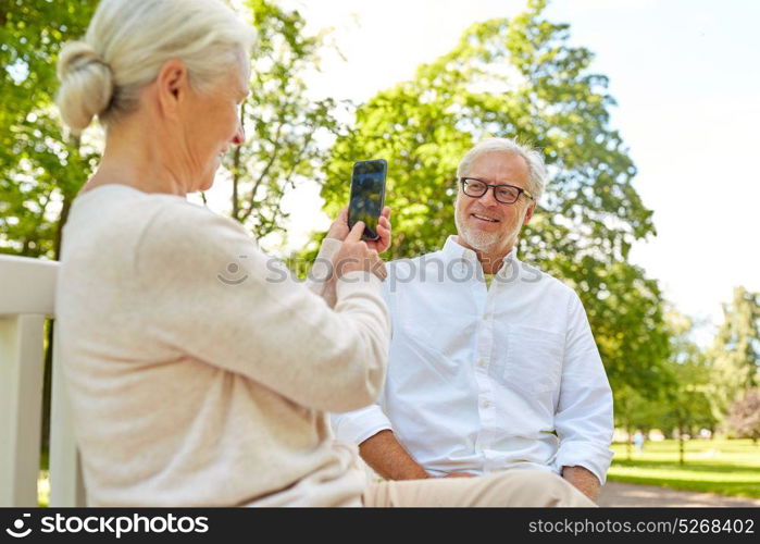 technology, relationship and old people concept - happy smiling senior couple with smartphone photographing in summer park. old woman photographing man by smartphone in park