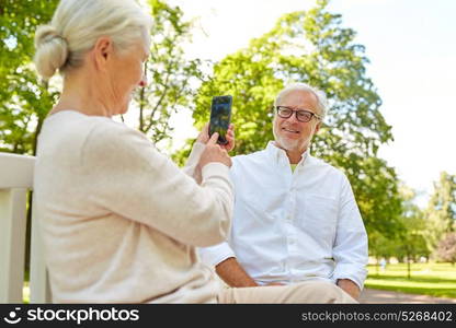 technology, relationship and old people concept - happy smiling senior couple with smartphone photographing in summer park. old woman photographing man by smartphone in park