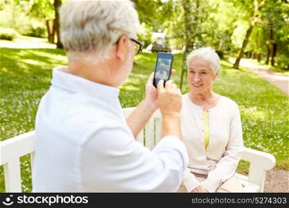 technology, relationship and old people concept - happy smiling senior couple with smartphone photographing in summer park. old man photographing woman by smartphone in park