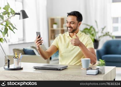 technology, remote job and gesture concept - happy smiling indian man with smartphone and laptop computer having video call and showing thumbs up at home office. indian man having video call on smartphone at home