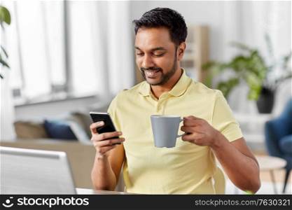 technology, remote job and lifestyle concept - happy indian man with smartphone drinking coffee or tea at home office. man with smartphone drinking coffee at home office