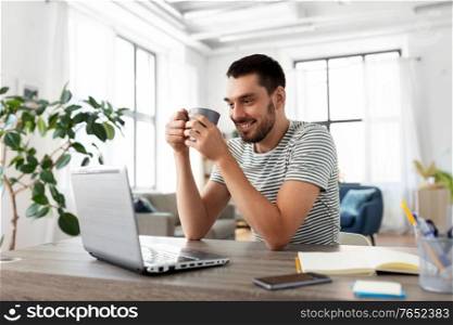 technology, remote job and lifestyle concept - happy man with laptop computer drinking coffee or tea at home office. man with laptop drinking coffee at home office