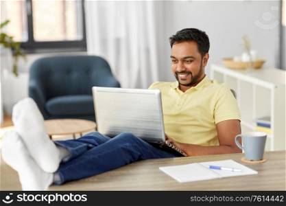 technology, remote job and lifestyle concept - happy smiling indian man with laptop computer resting feet on table at home office. man with laptop resting feet on table at home