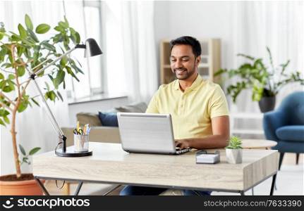 technology, remote job and lifestyle concept - happy smiling indian man with laptop computer working at home office. indian man with laptop working at home office