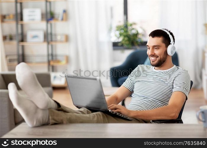 technology, remote job and lifestyle concept - happy smiling man with laptop computer and headphones resting feet on table at home office. happy man with laptop and headphones at home