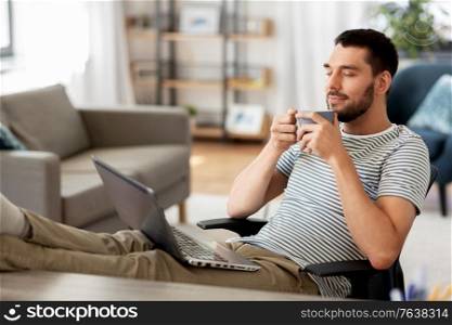 technology, remote job and lifestyle concept - happy smiling man with laptop computer drinking coffee and resting feet on table at home office. happy man with laptop drinking coffee at home