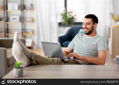 technology, remote job and lifestyle concept - happy smiling man with laptop computer and earphones resting feet on table at home office. happy man with laptop and earphones at home