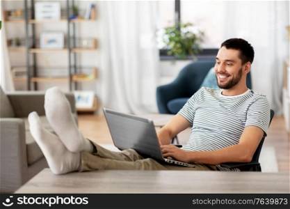 technology, remote job and lifestyle concept - happy smiling man with laptop computer resting feet on table at home office. man with laptop resting feet on table at home