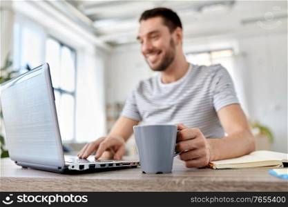 technology, remote job and lifestyle concept - happy smiling man with laptop computer drinking coffee at home office. happy man with laptop drinking coffee at home