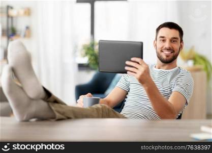 technology, remote job and lifestyle concept - happy smiling man with tablet pc computer resting feet on table at home office. man with tablet pc resting feet on table at home