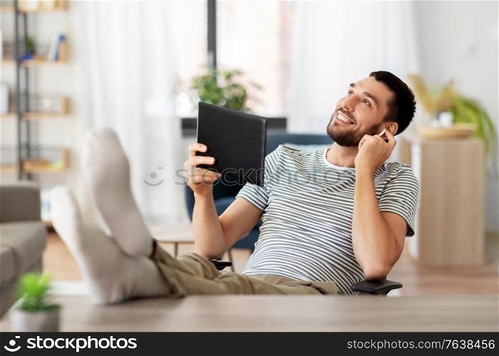 technology, remote job and lifestyle concept - happy smiling man with tablet pc computer and earphones resting feet on table at home office. happy man with tablet pc and earphones at home