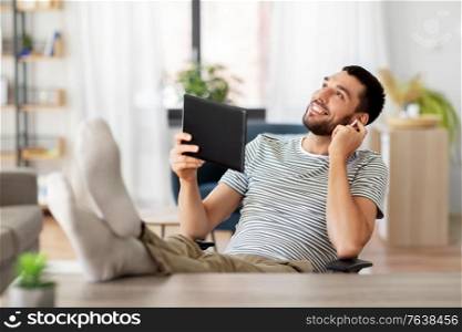 technology, remote job and lifestyle concept - happy smiling man with tablet pc computer and earphones resting feet on table at home office. happy man with tablet pc and earphones at home