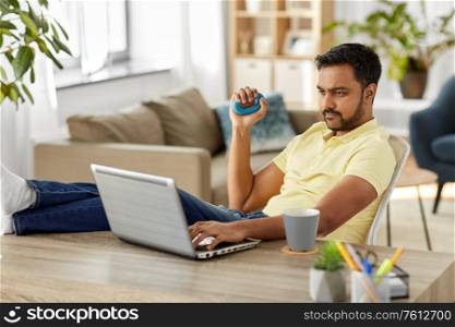 technology, remote job and lifestyle concept - indian man with laptop computer gripping hand expander and resting feet on table at home office. man with laptop and hand expander at home office